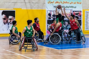 بازیکنان که پیراهن قرمز به تن دارند در حال دفاع از پرتاب توپ به سمت حلقه بازیکن تیم مقابل هستند.